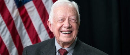President Jimmy Carter in 2014 smiles in front of an American flag set against a black background