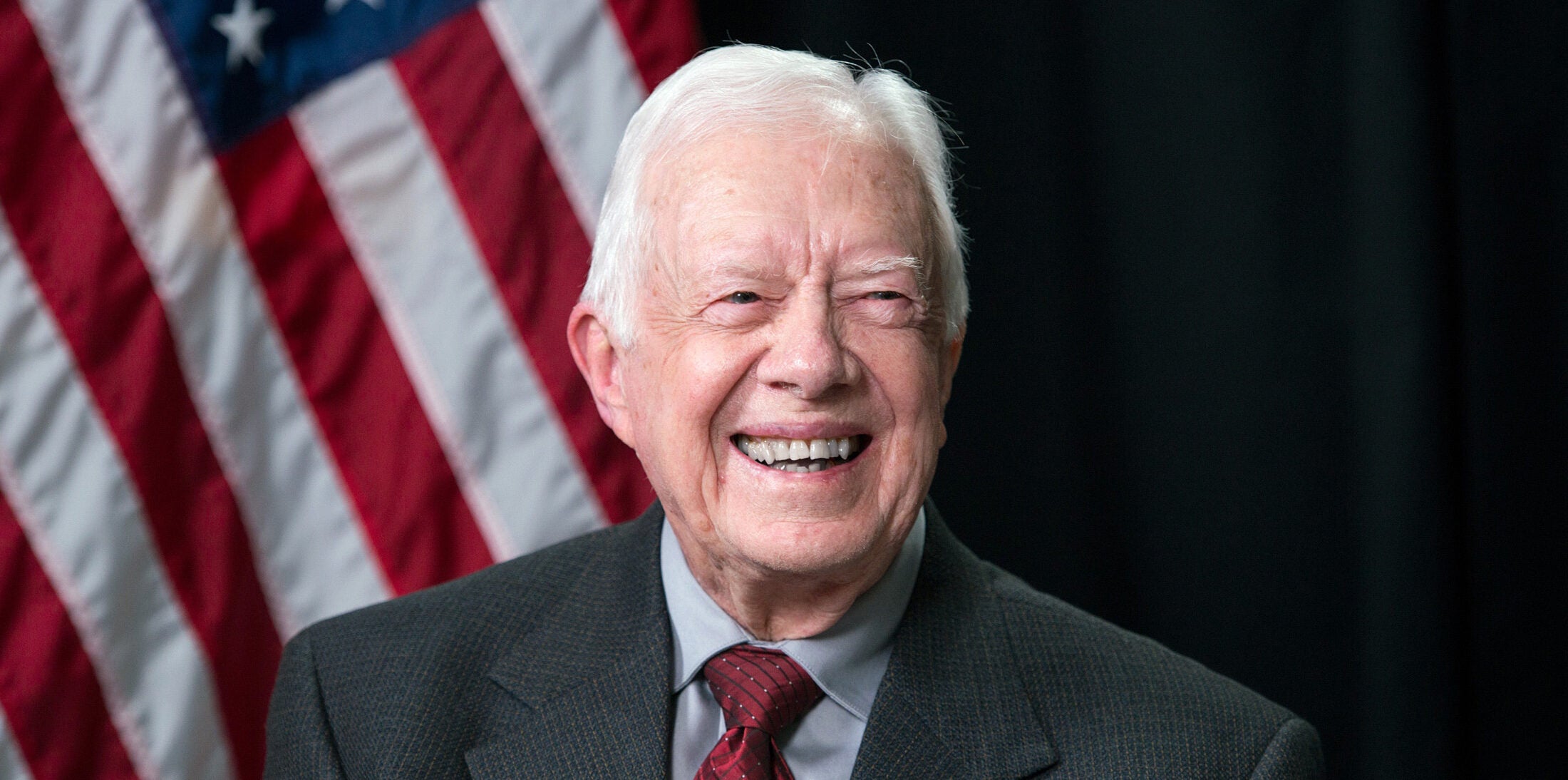 President Jimmy Carter in 2014 smiles in front of an American flag set against a black background